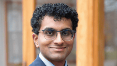 Man with dark curly hair wearing glasses and a suit