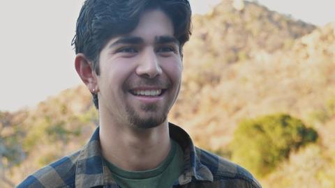 Man with dark hair wearing plaid shirt and standing in front of a mountain