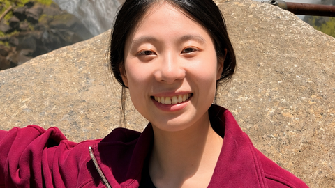 Portrait of PhD candidate Jingting Liang standing outside by a waterfall wearing a red sweater