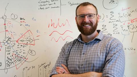 Man with beard and glasses wearing a plaid shirt standing in front of a white board with writing on it