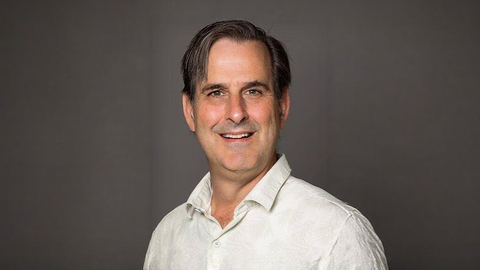 Man with dark hair wearing a white shirt and smiling in front of a dark gray background