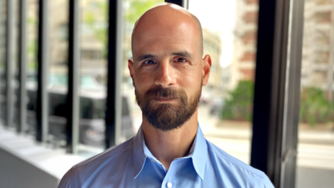 Man with beard wearing a blue shirt standing in front of windows