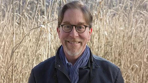 Man wearing glasses, a black jacket and scarf standing in a field 