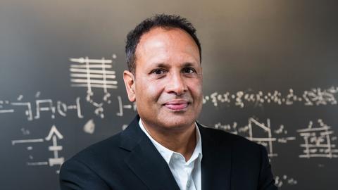 Man with dark hair wearing a black suit and standing in front of a chalk board with writing