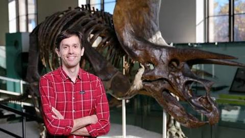 Man with crossed arms wearing a red checked shirt standing in front of a fossil