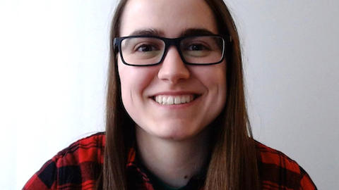 Catherine Strong with long brown hair, waring glasses and a red and black flannel top, in front of a light grey background