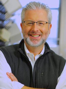 Man with beard and glasses wearing black vest and white shirt smiling