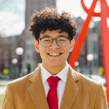 Man with curly hair and glasses wearing a tan suit standing outside