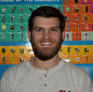 Man with a beard wearing a white top standing in front of a poster