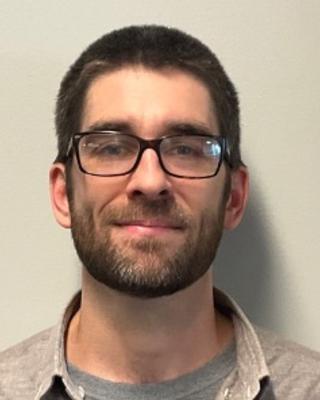 Peter D Wiggan with short brown hair and beard, grey t-shirt, grey button down, light grey background