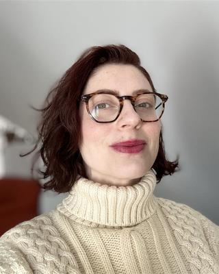 Jenna Legault with auburn hair, tortoiseshell glasses, turtleneck cream sweater, blurred room background