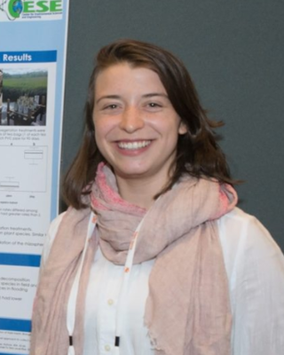 Alaina Bisson with long brown hair, pale pink scarf, and whit shirt, in front of scientific poster and grey wall