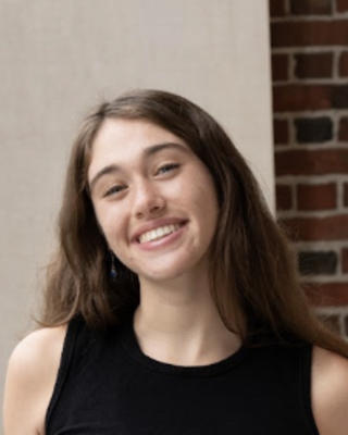 Beatrice Youd with long brown hair and black sleeveless top, in front of cream wall and brick column