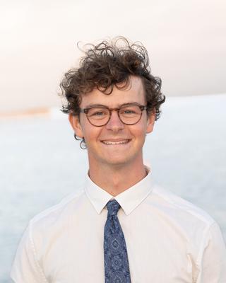 Max Laubstein with short curly brown hair, wearing glasses and a white shirt and blue tie, with the ocean in the background