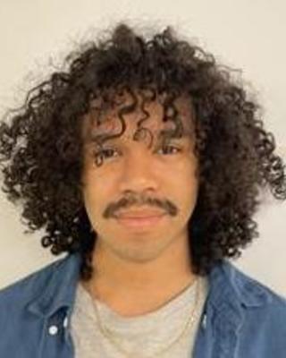 Melvin Williams with long curly cark hair, wearing grey top and blue top over it, with beige background
