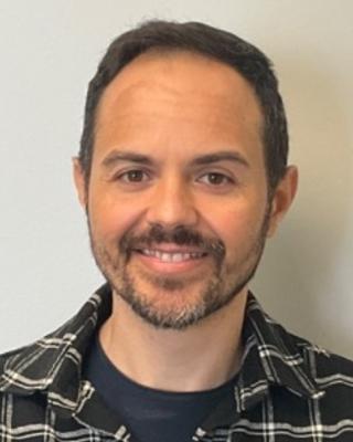 Paulino Siqueira Ribeiro with short brown hair and beard, wearing dark plaid shirt, in front of light grey background