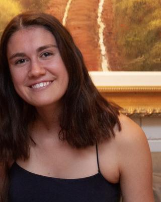 Ella Frigyik with long brown hair and a dark top, in front of a warm-toned painting