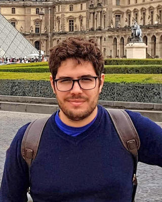 Bruno Kerber with short brown hair and dark glasses, wearing blue sweater, in front of the Louvre