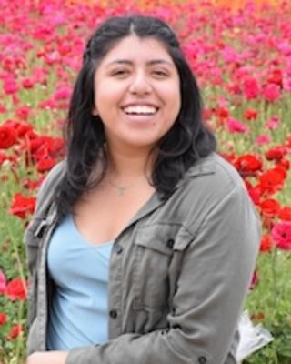 Adriana Aguilar-Maldonado with long dark hair, blue top and grey jacket, in a field of red flowers