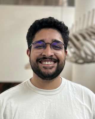 Akshay Kan with short black hair and beard, wearing glasses and a white shirt, with fossil in background