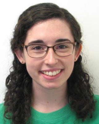 Emily Rivard with long curly brown hair, in a green top wearing glasses, in front of a white background