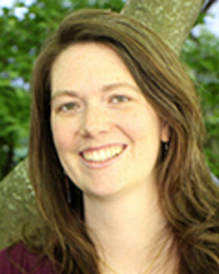 Kelsey Tyssowski with long brown hair, wearing a maroon shirt, in front of a tree and shrubs