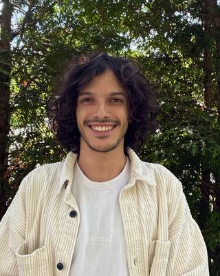Lucas Funaro with long dark wavy hair, a white shirt and cream corduroy shirt, in front of green trees