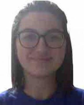 Maddi Waskom with long dark hair, wearing glasses and a blue top, in front of a white background