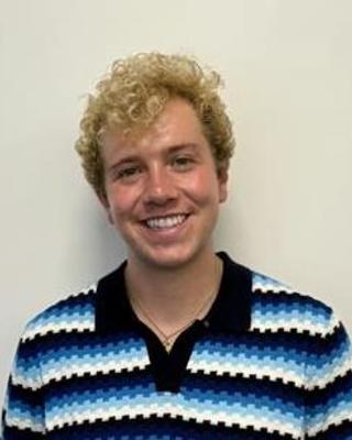 Seth Thomas with short blonde curly hair and blue striped top, in front of a beige background