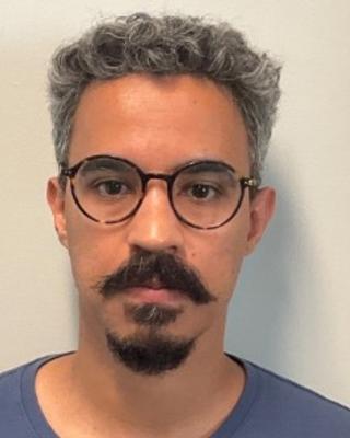 Williams L Porto with gray curly hair, glasses, and a dark mustache and beard, beige background