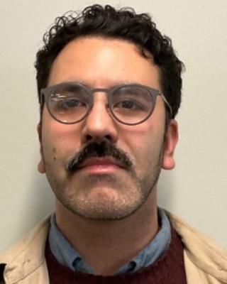 Alonso Delgado with dark, curly hair, mustache and glasses wearing a blue shirt, red sweater and tan jacket