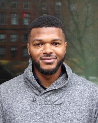 David Brewster with short black hair and beard wearing a gray shirt, in front of window reflecting trees and brick building