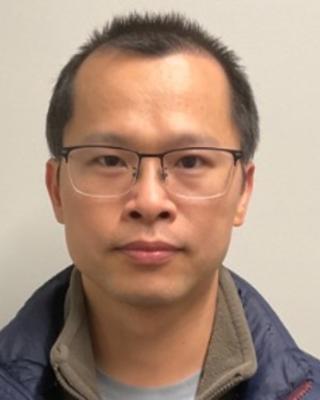 Bifeng Wu with short dark hair, wearing glasses and a blue coat, in front of a beige background