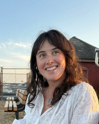 Julia Lucey, woman with long dark hair sitting outside wearing a white top