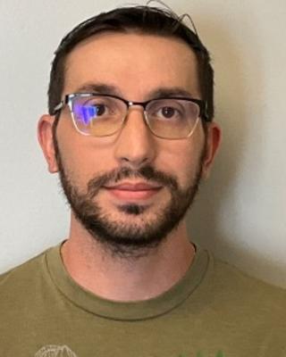 Portrait of postdoc Nikolaos Polizos wearing glasses, beard, and a green shirt