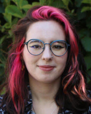 Portrait of graduate student Ana Stratan with long hair with pink streaks wearing glasses and standing outside