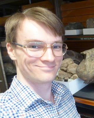 Portrait of postdoc Peter Bishop standing in front of a fossil wearing a plaid shirt and glasses
