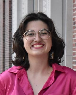 Portrait of PhD candidate maddi Waskom standing in front of a brick building wearing a pink top and glasses