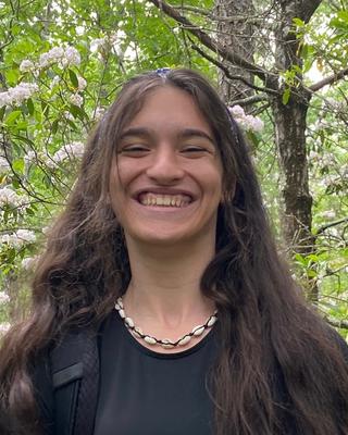Woman with long dark hair wearing a black top and necklace, smiling in front of trees
