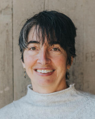 Alex Worden with shot dark hair, wearing a white sweater in front of a light wood background