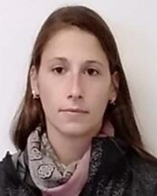 Amandine Gillet with long brown hair, pink scarf, and dark coat, in front of beige background