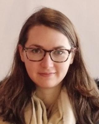 Andrea Berardi with long brown hair, glasses, cream sweater and dark coat, in front of beige background