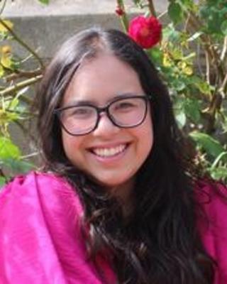 Andrea Loya Castillo with long dark hair, glasses, and a bright pink top, in front of dark pink rose bushes