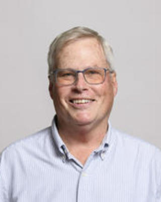 Andrew Biewener smiling, with light grey hair, light blue shirt, in front of grey background