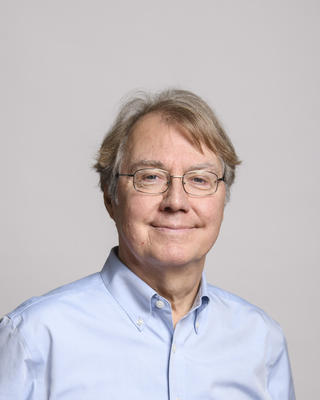 Andrew H. Knoll with short light hair, wearing a light blue button down and glasses, smiling, with grey background