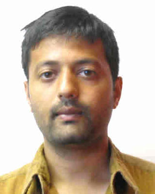 Ashesh Dhawale with short black hair, wearing a dark yellow shirt, in front o flight grey background