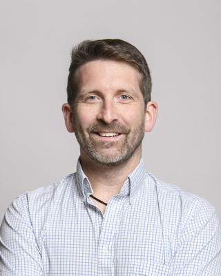 Benton Taylor with dark hair and a beard wearing a white shirt, in front of grey background