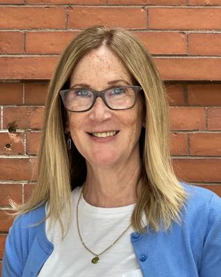 Bridget Power, smiling, with long hair, white shirt, light blue cardigan, brown glasses, in front of brick wall