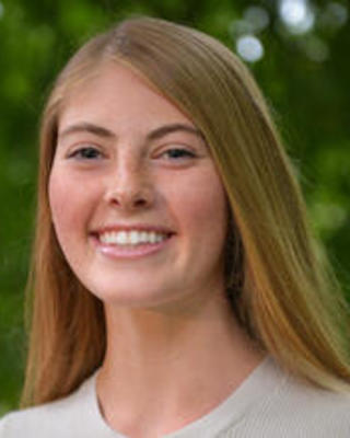 Brooke Travis with long blonde hair, wearing a white shirt, leafy trees in background