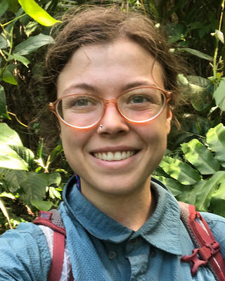 Carina Berlingeri with brown hair pulled back, glasses, blue shirt, and red backpack, surrounded by green leaves